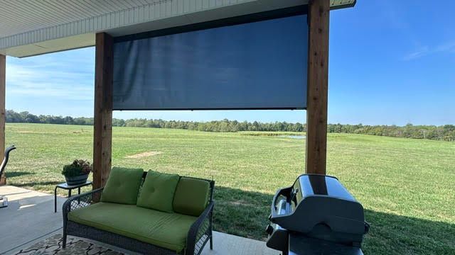 Outdoor patio with lowered black sun shade, green couch, grill, overlooking a field.