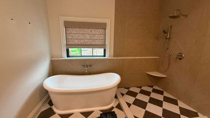 Bathroom with white tub, shower, and patterned floor. Window with shade. Light tan walls.