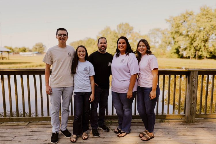 Family of five poses on a wooden deck overlooking water. All wear casual clothes, smiling at the camera.