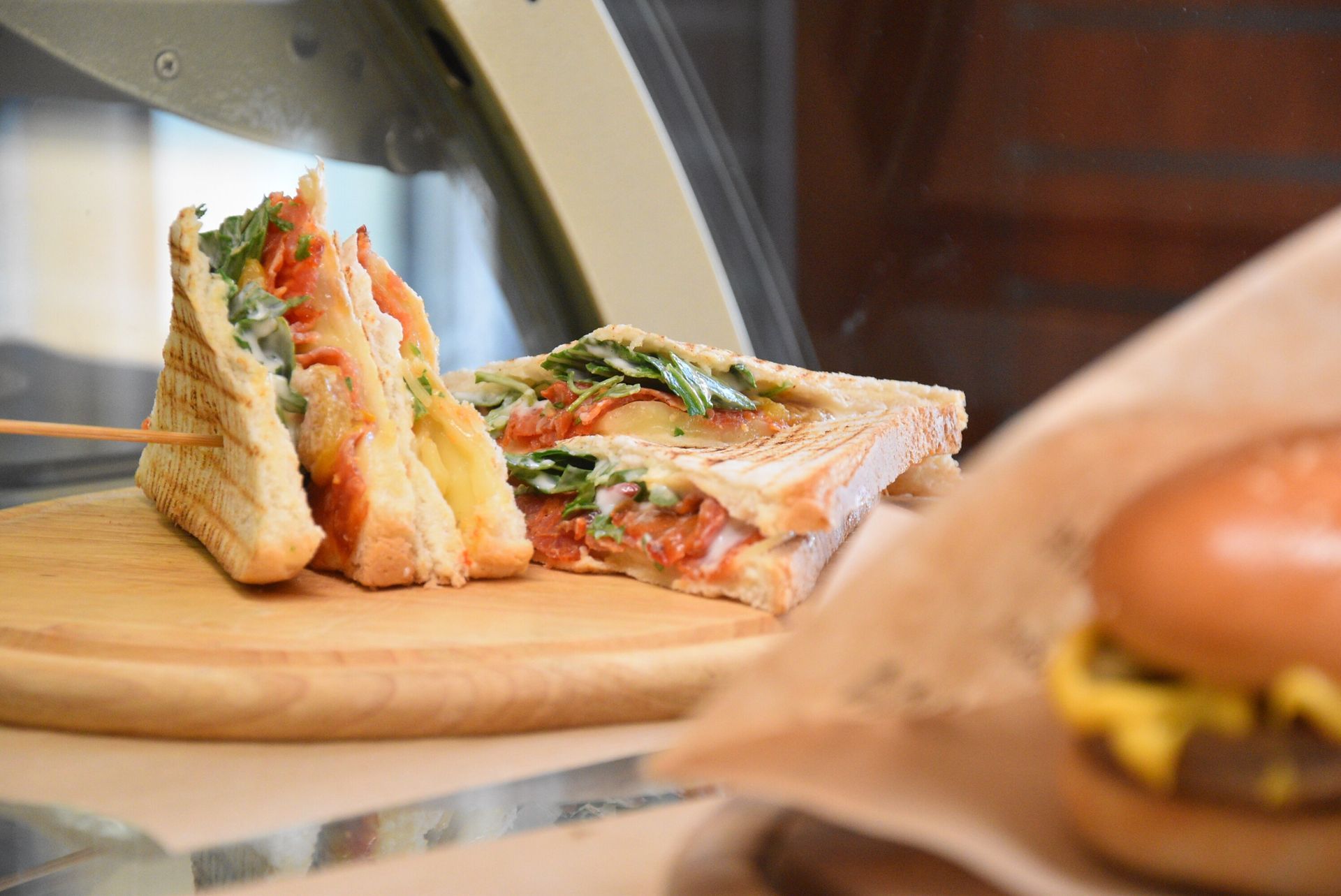 Club sandwich tostato su un tagliere di legno con hamburger in primo piano.