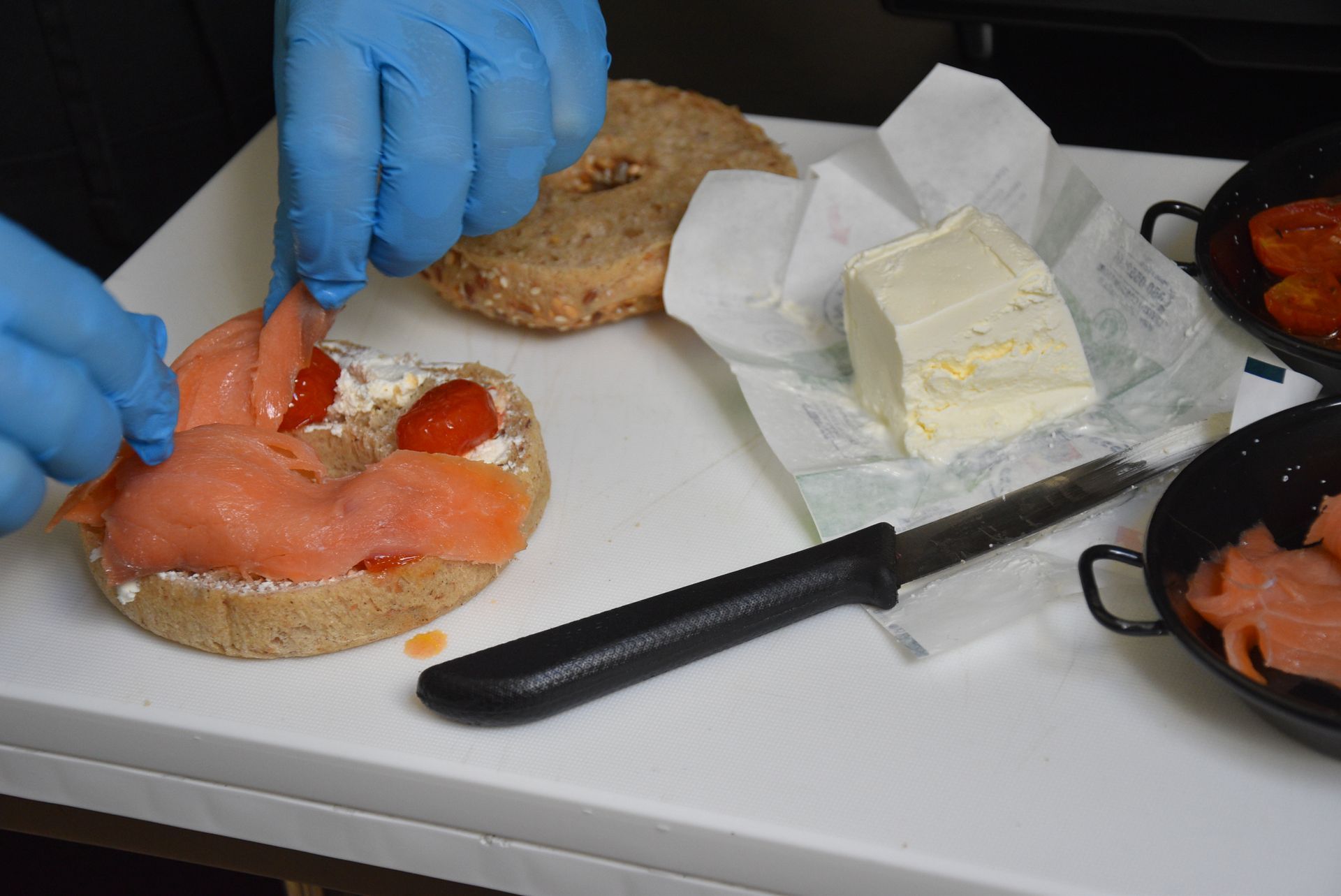 Mani con guanti blu che appoggiano il salmone affumicato su un bagel con formaggio cremoso, tagliere bianco.