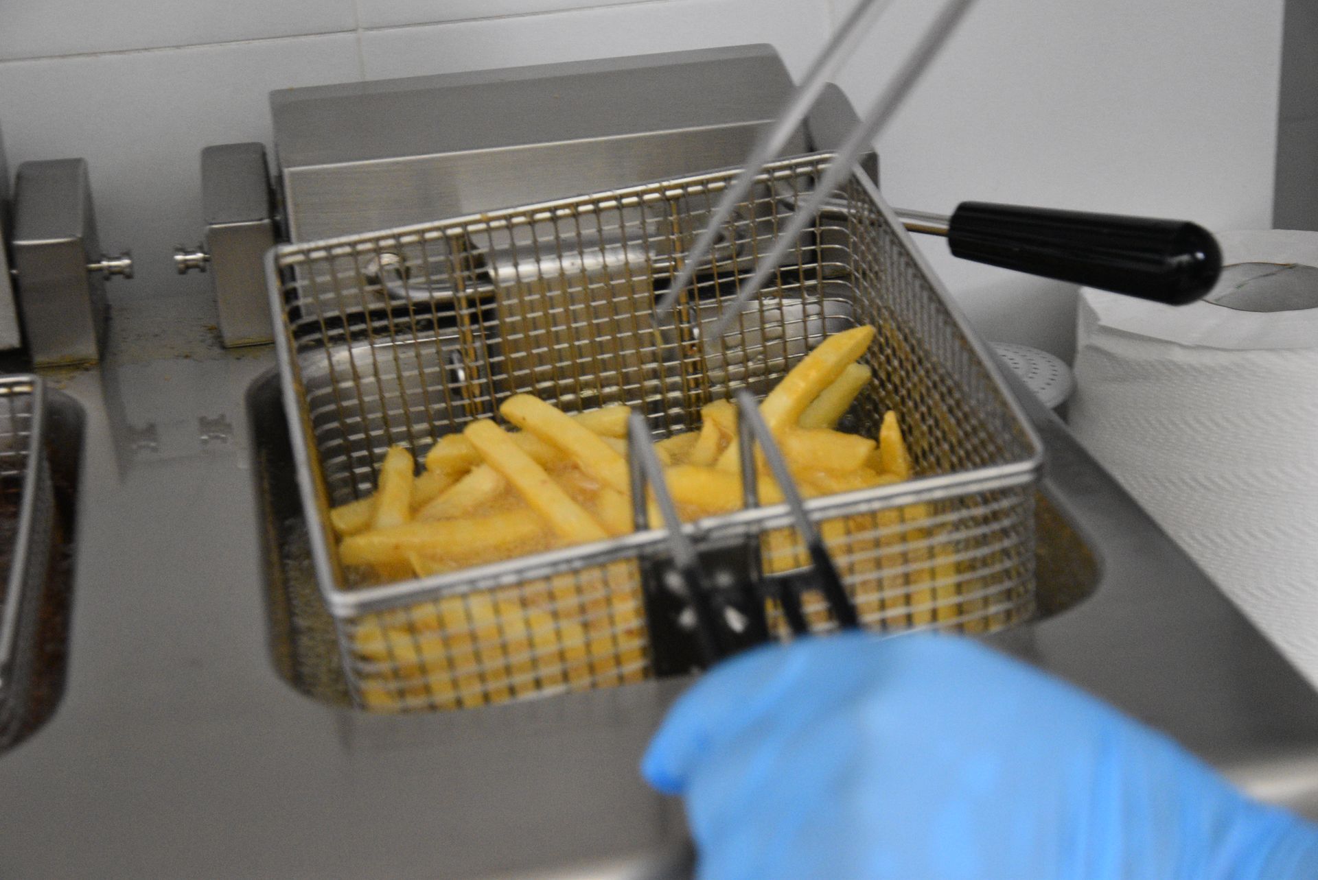 Una persona con un guanto blu solleva un cestino di patatine fritte da una friggitrice con le pinze.