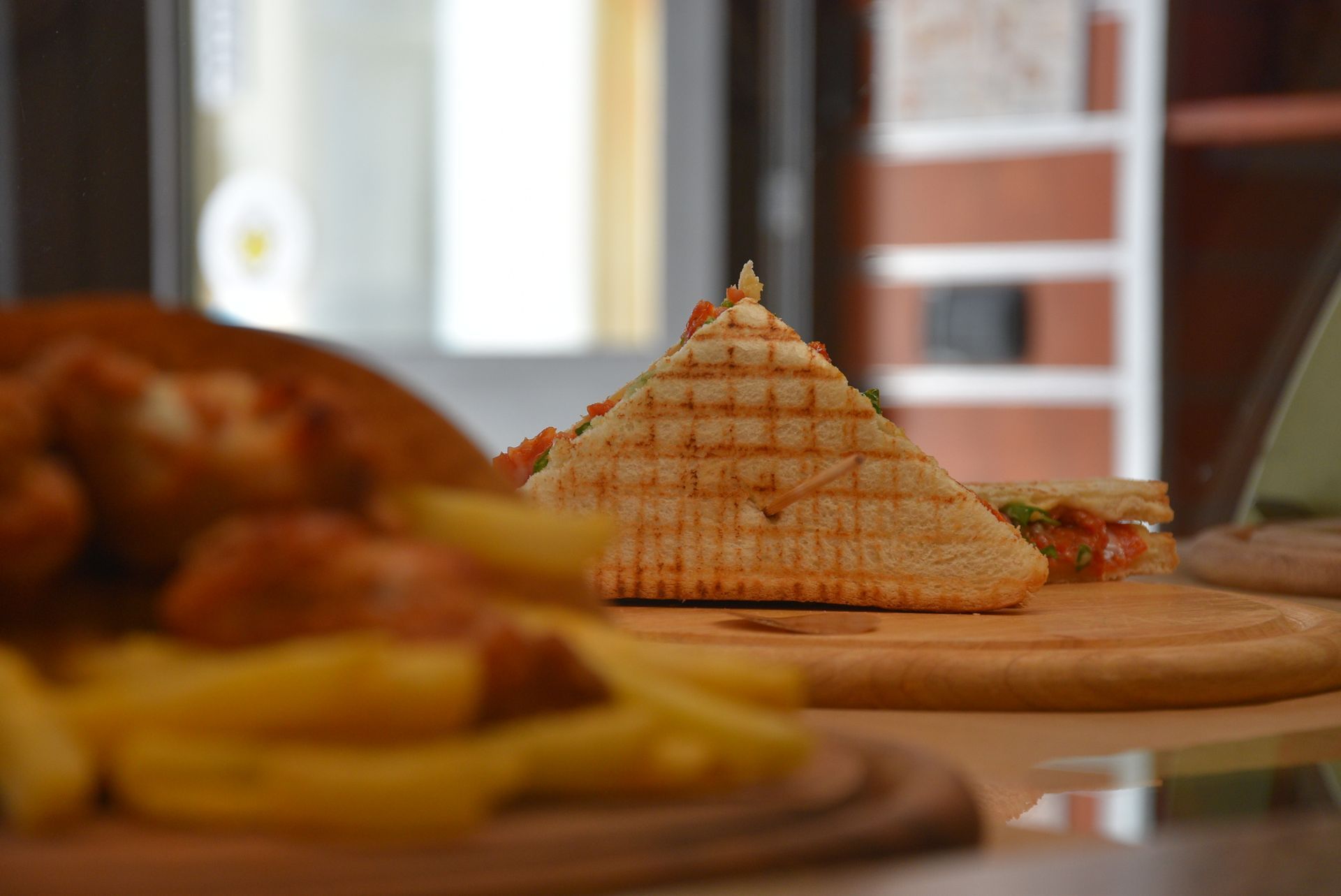 Panino grigliato su un tagliere di legno, con patatine fritte e cibo fritto sfocati in primo piano.