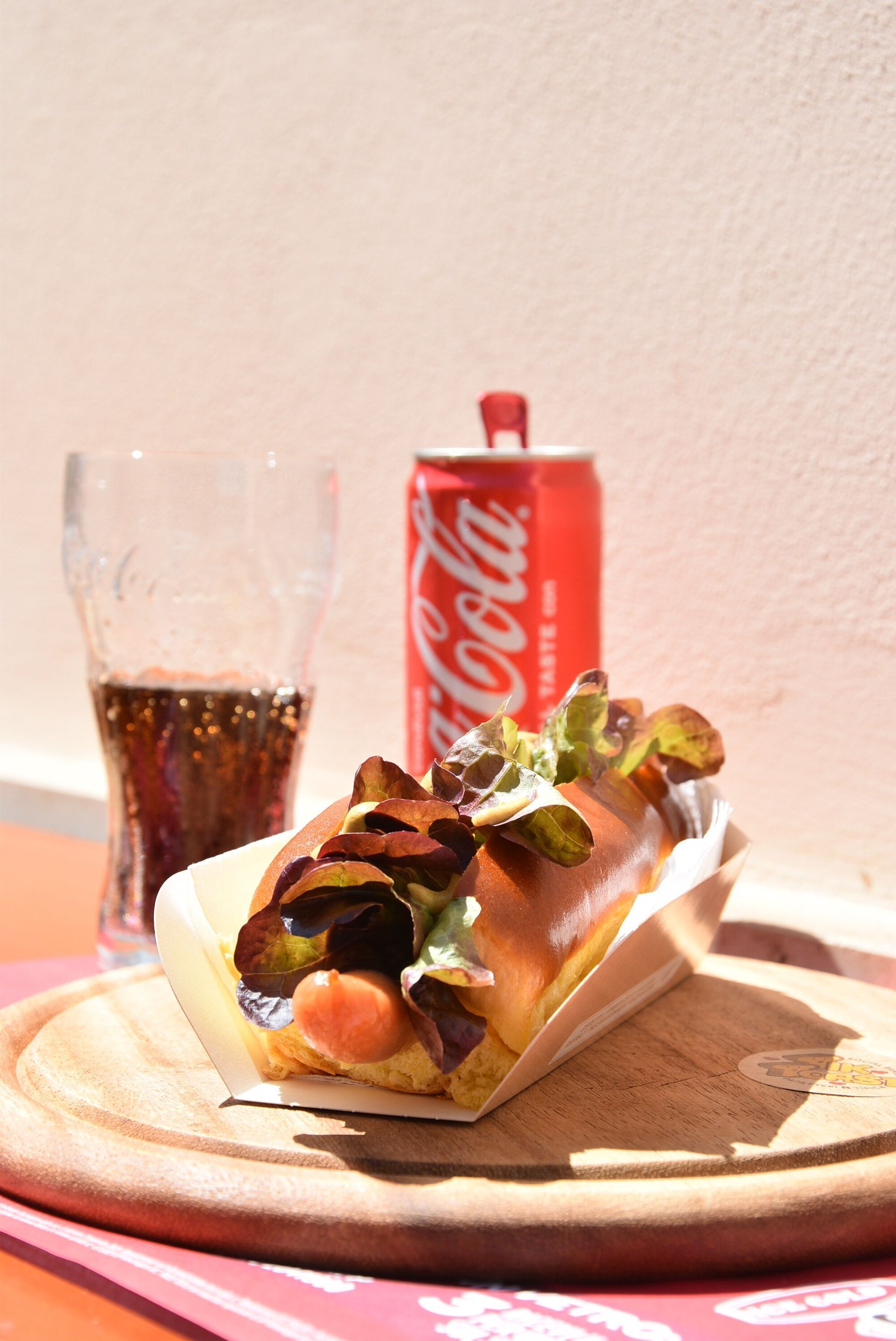 Hot dog con lattuga e bibita, lattina di Coca-Cola e un bicchiere di soda su un piatto di legno.