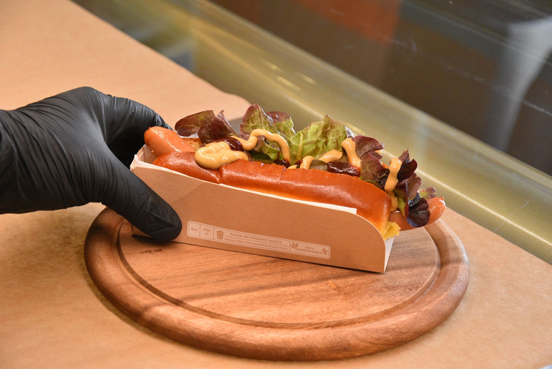 Un hot dog in un vassoio di carta, guarnito con senape e lattuga, tenuto da una mano guantata su un piatto di legno.