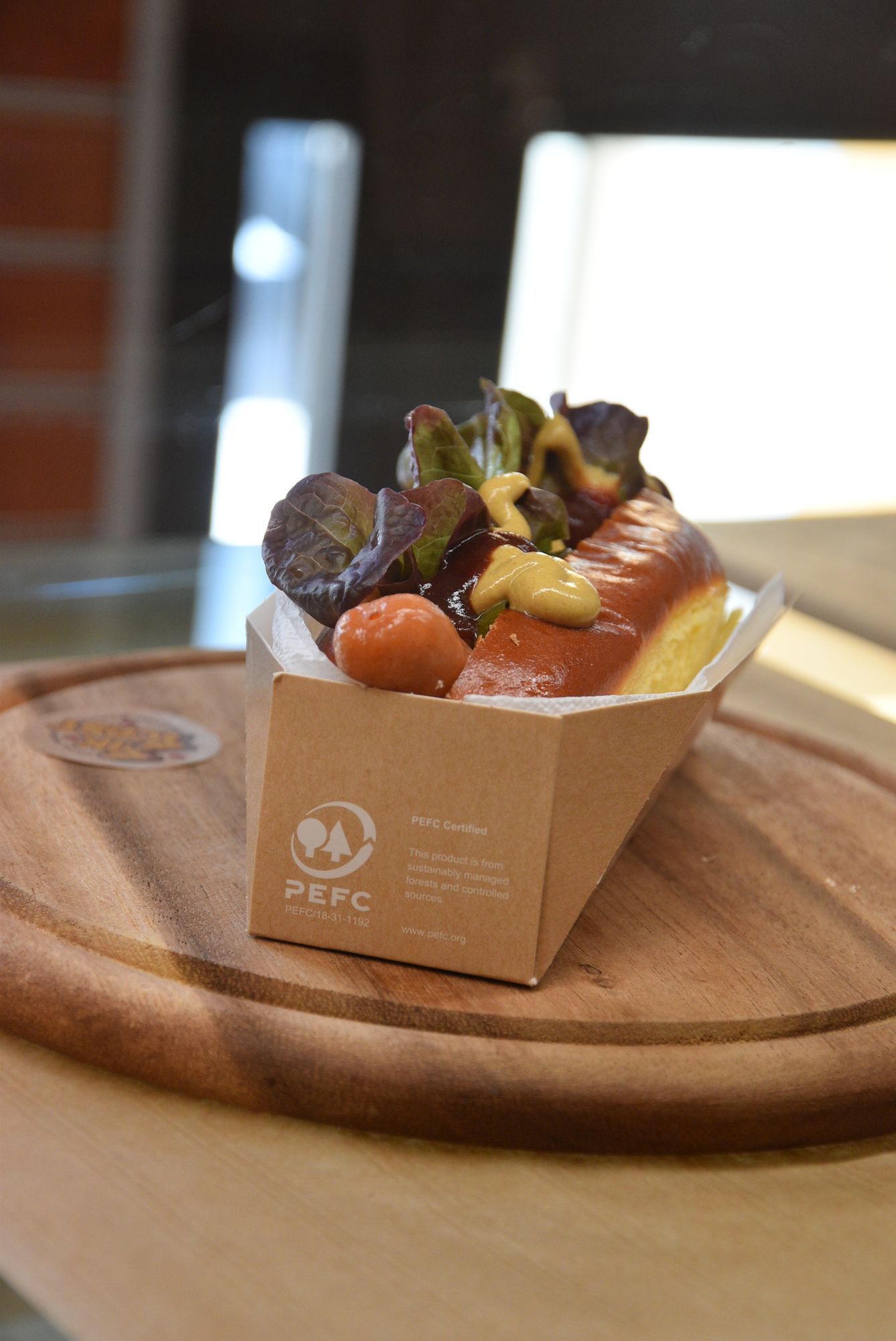 Hot dog in un contenitore di cartone su un vassoio di legno, guarnito con verdure e senape.