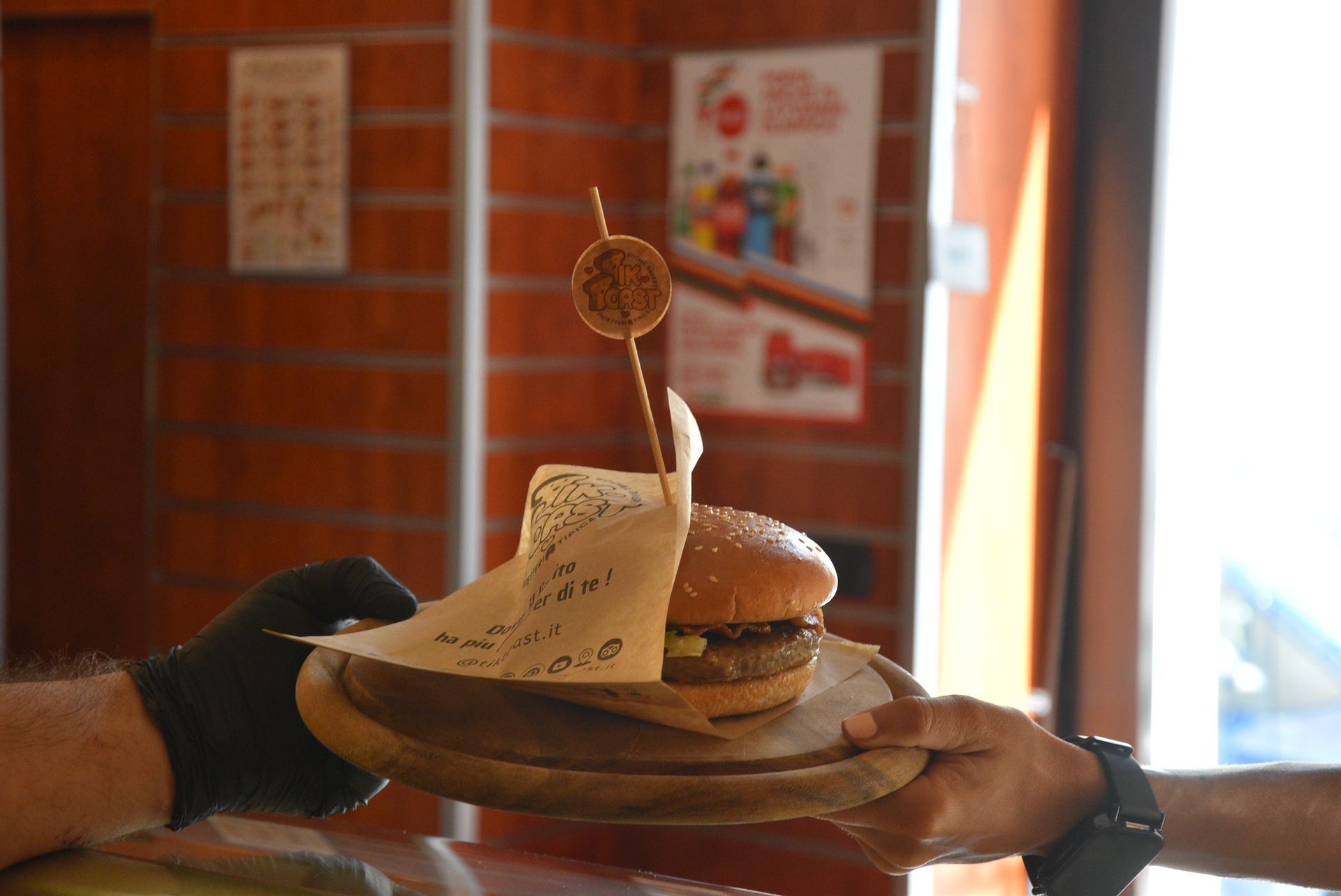 Hamburger su un piatto di legno, tenuto da mani guantate. Uno spiedino decorativo sostiene una polpetta fritta.