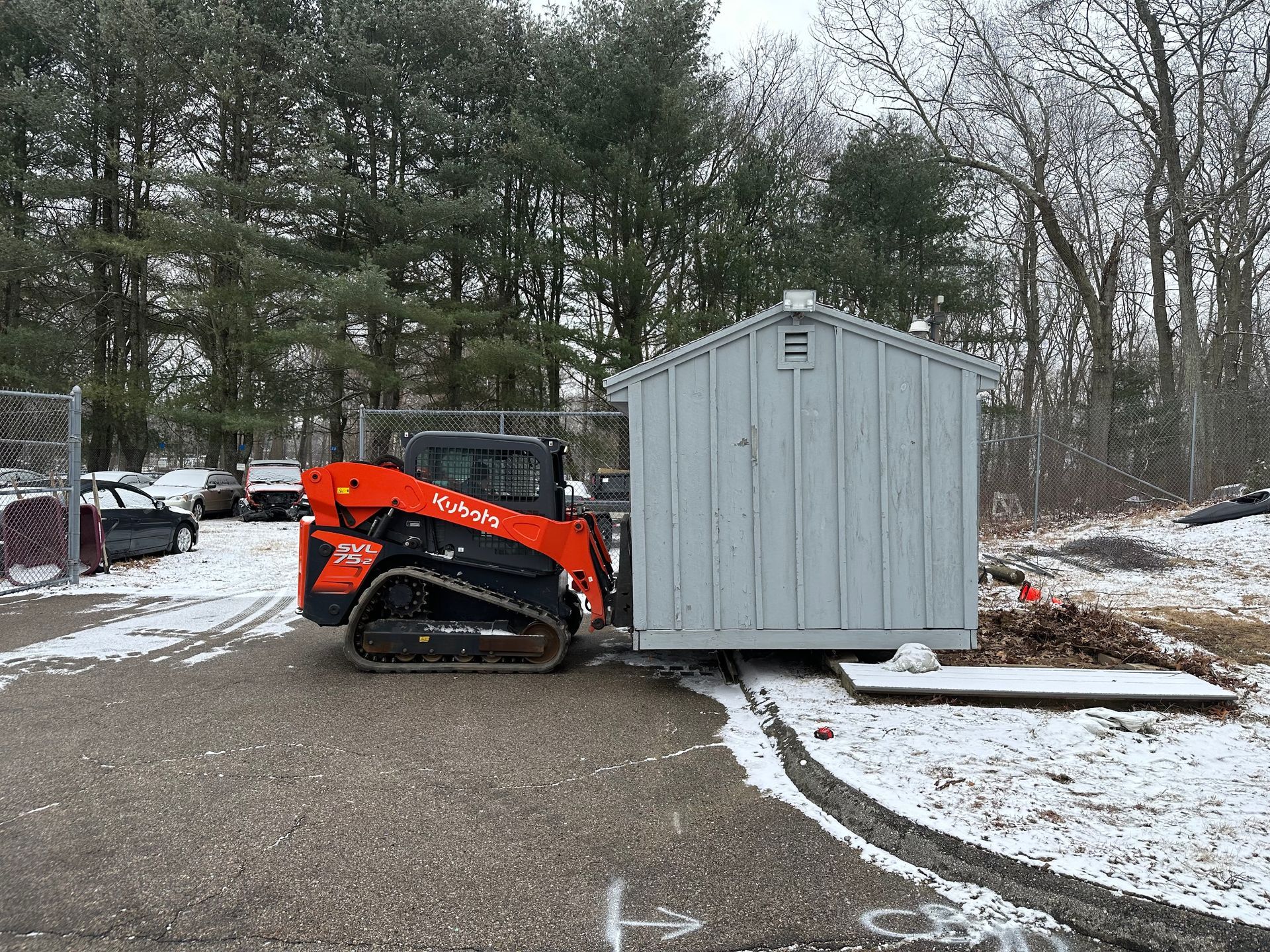 Orange skid steer moving a light blue shed on a snowy asphalt surface, trees in the background.