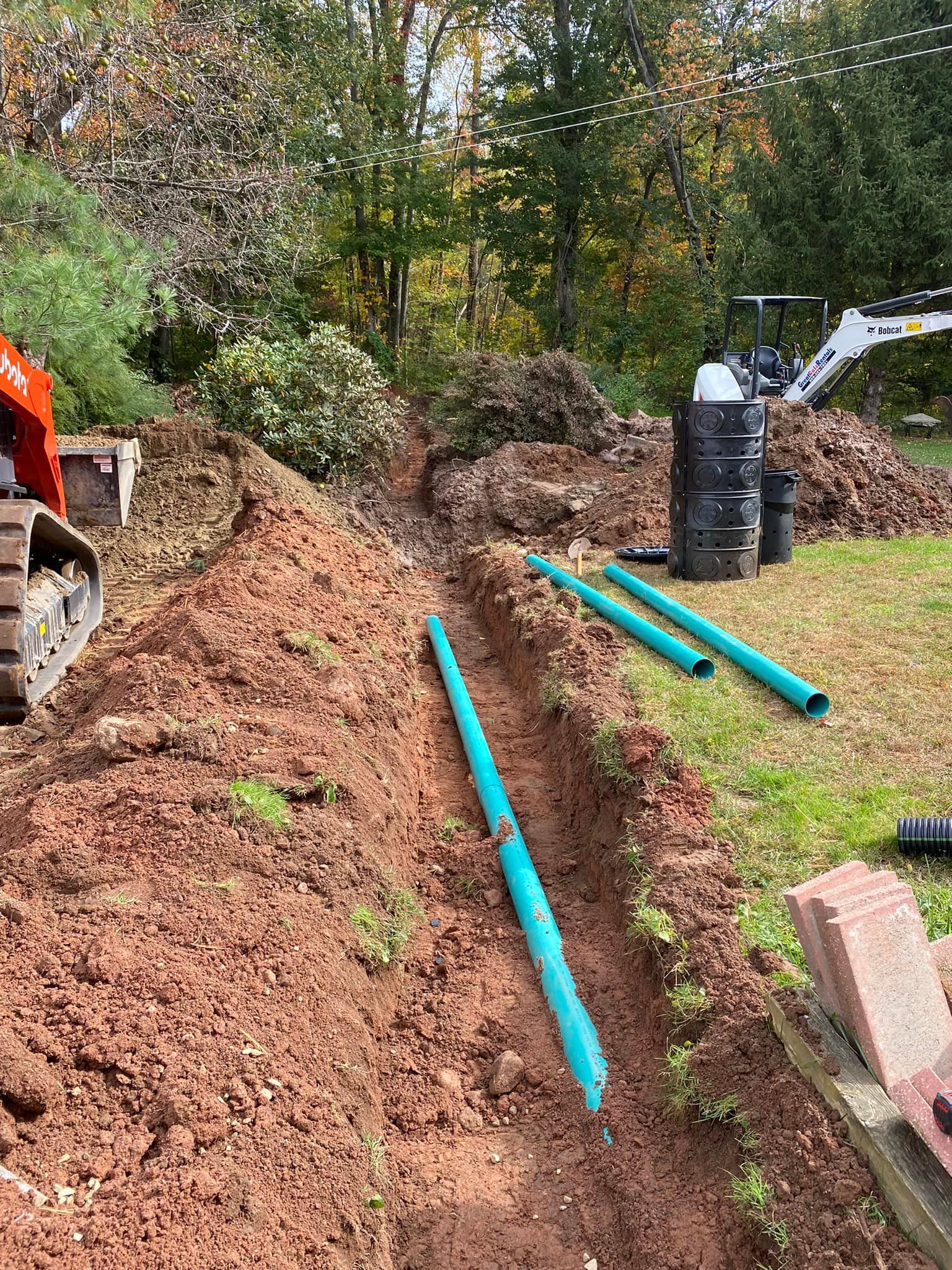 Trench dug in soil with turquoise drainage pipes. Excavator and trees in the background.