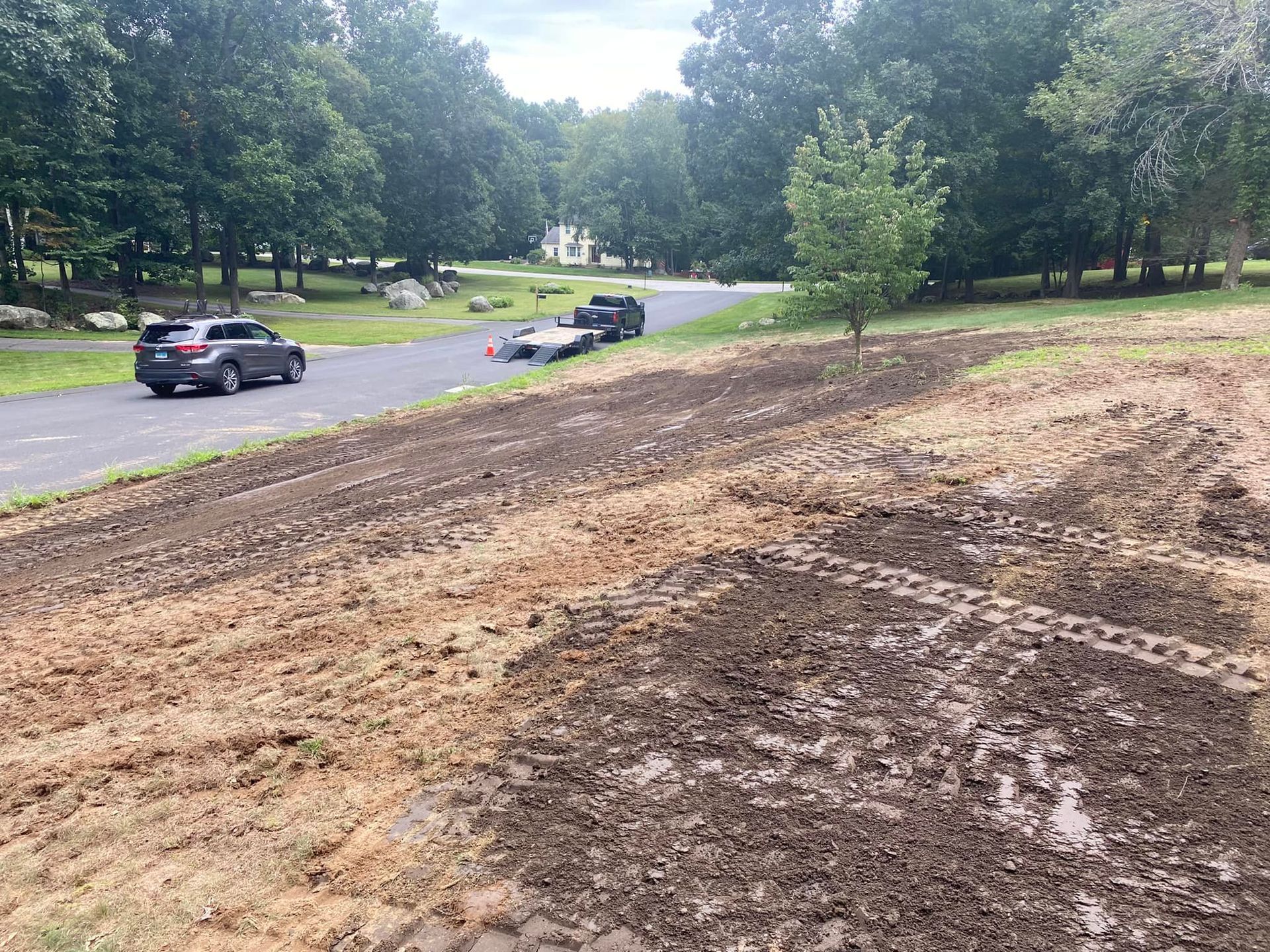 Dirt yard with a road, a car, and a trailer. Trees line the background.