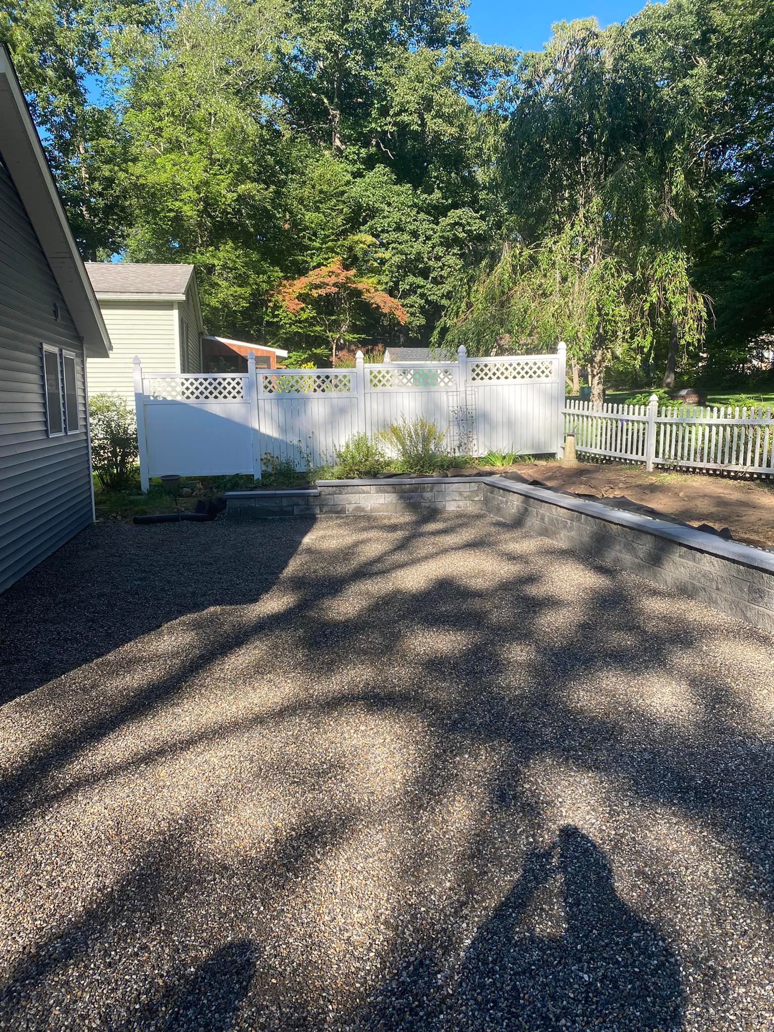 Gravel driveway beside a light-colored house. A white fence is in the background, with trees overhead.