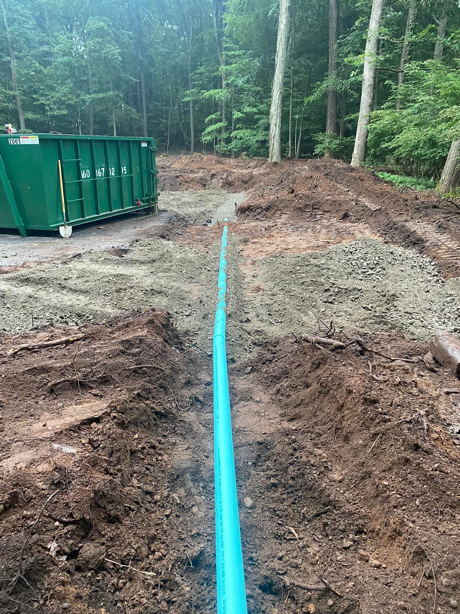 Trench dug for a blue pipe, with a green dumpster and a wooded backdrop. Muddy ground.