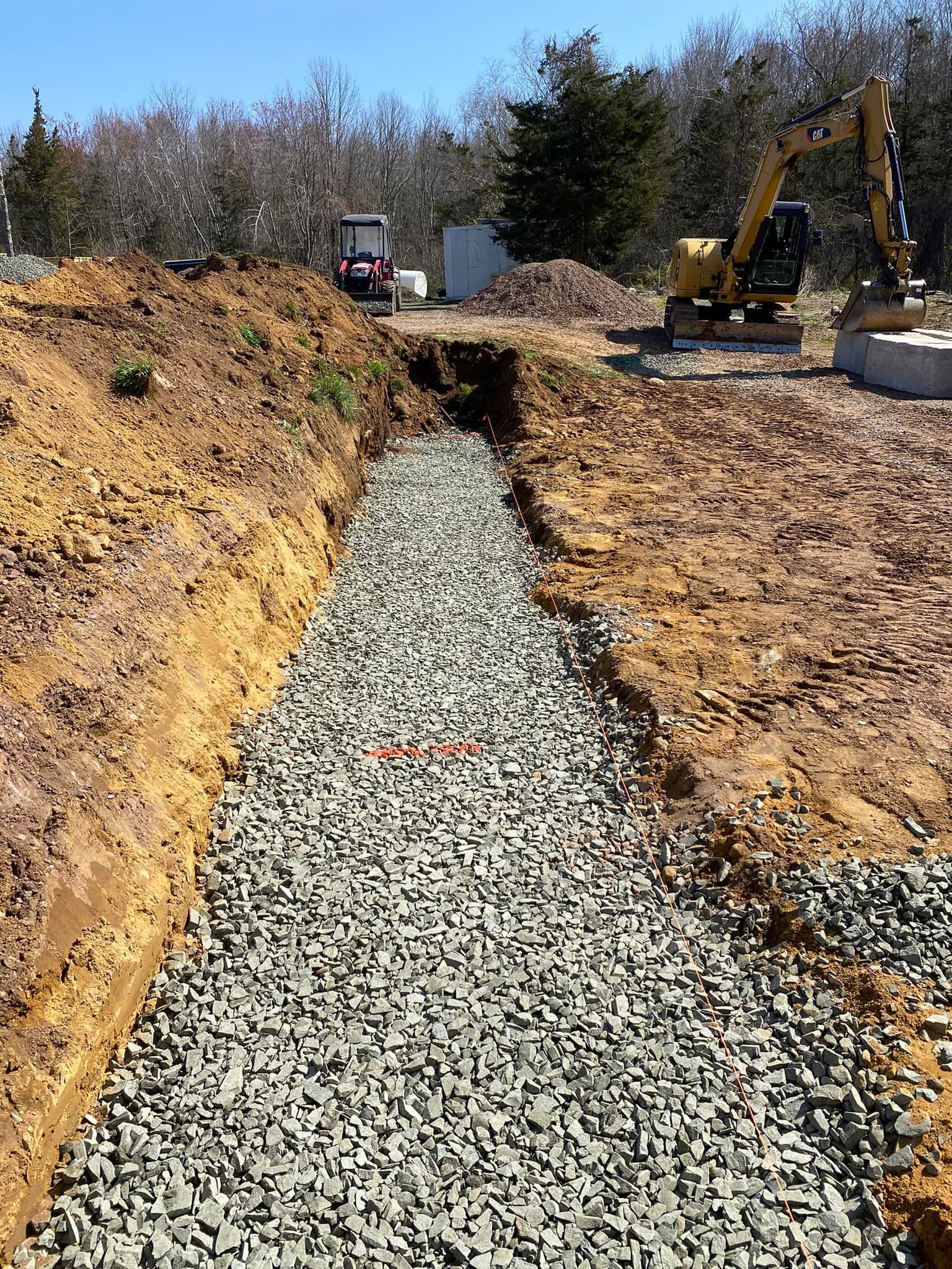 Construction site: Excavated trench filled with gravel. Yellow excavator and red tractor visible.