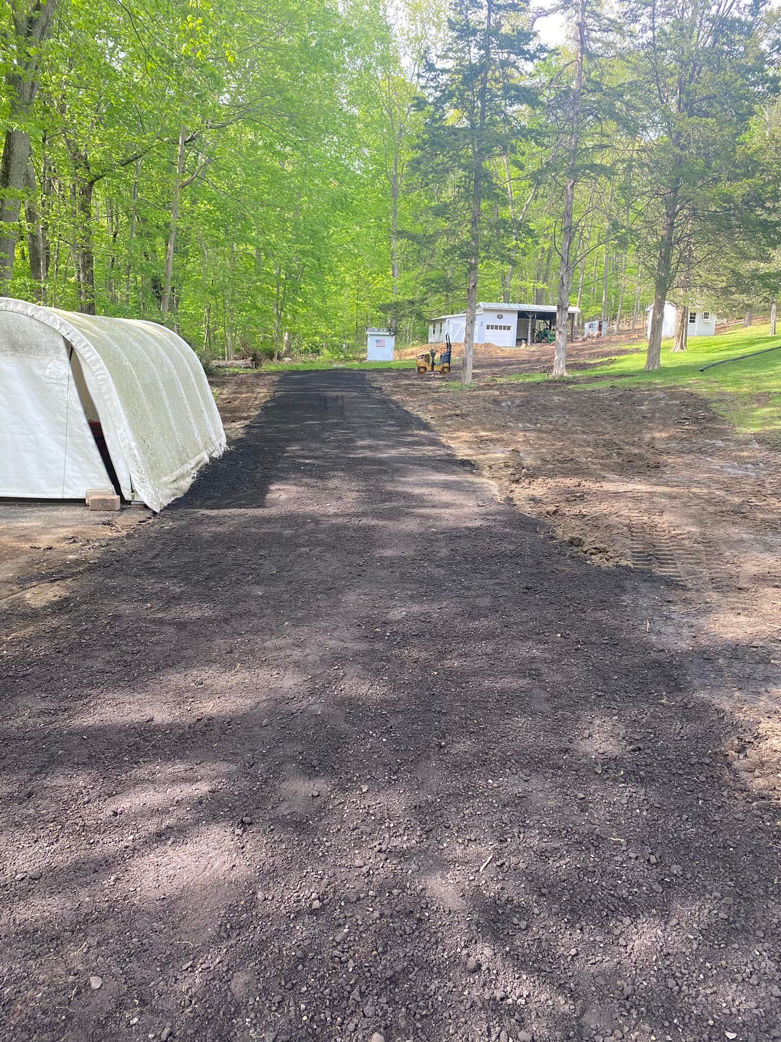 Long, dark mulch path leading to buildings in a rural area, with trees on both sides.