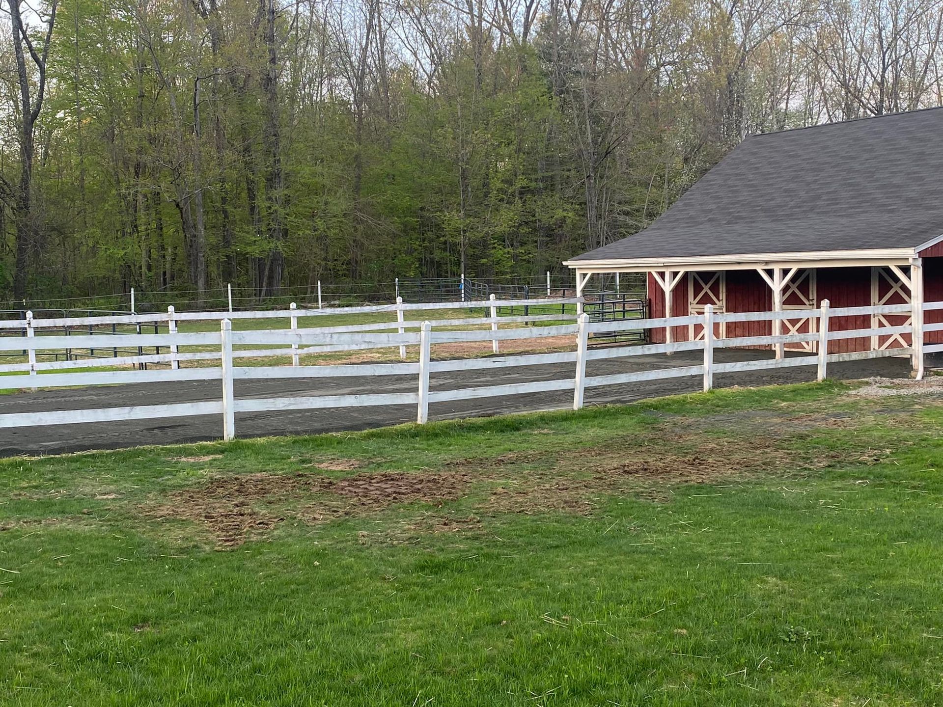 Red barn with white fencing and a gravel arena in a grassy area. Trees in background.
