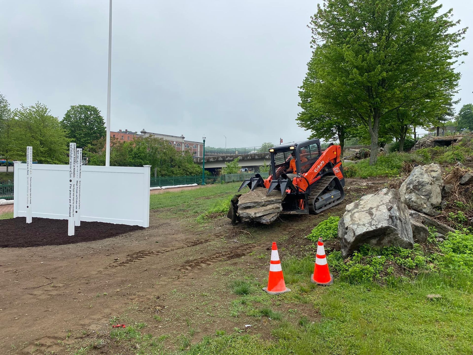 Orange skid steer on a hillside carrying a rock, near a white fence, under an overcast sky.