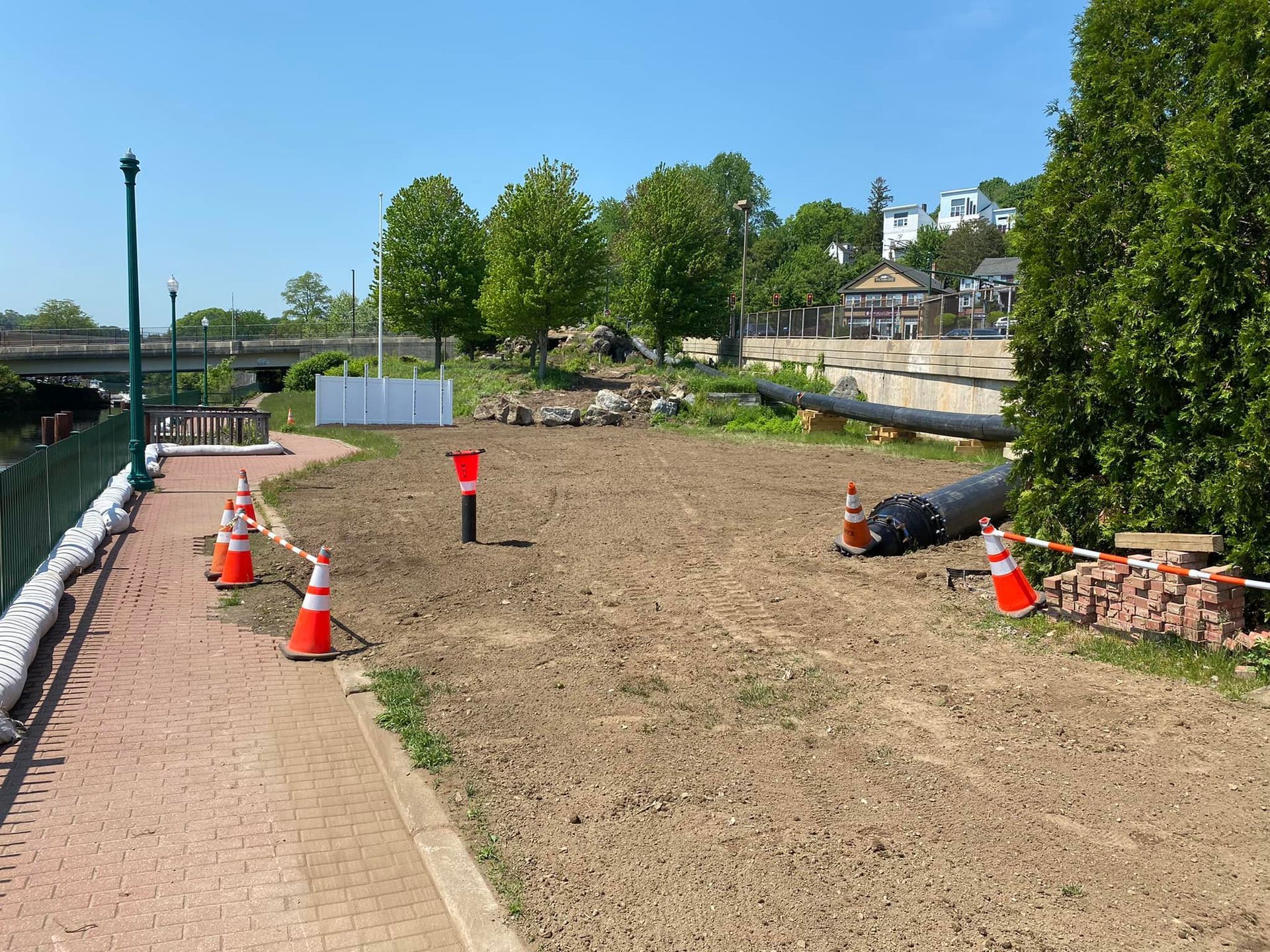 Construction zone with orange cones, dirt, and a black pipe. A pathway and trees are also visible.