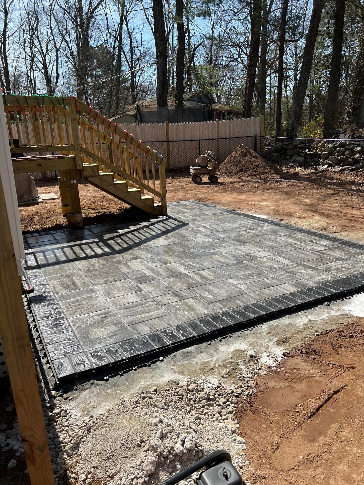 Wooden deck with stairs leads to a newly laid paver patio surrounded by dirt, in a wooded area.
