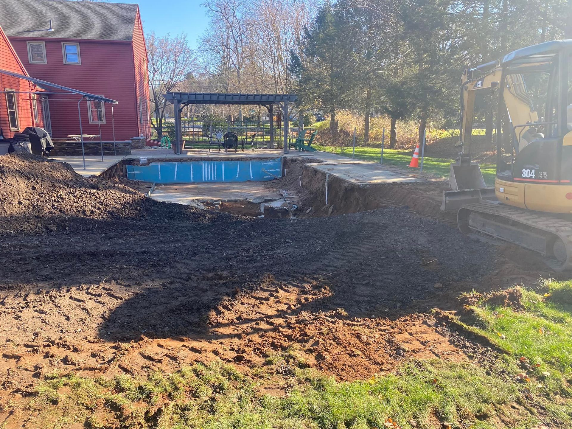 Excavation for a pool in a backyard, with an excavator and a house. Soil and a small pool are visible.