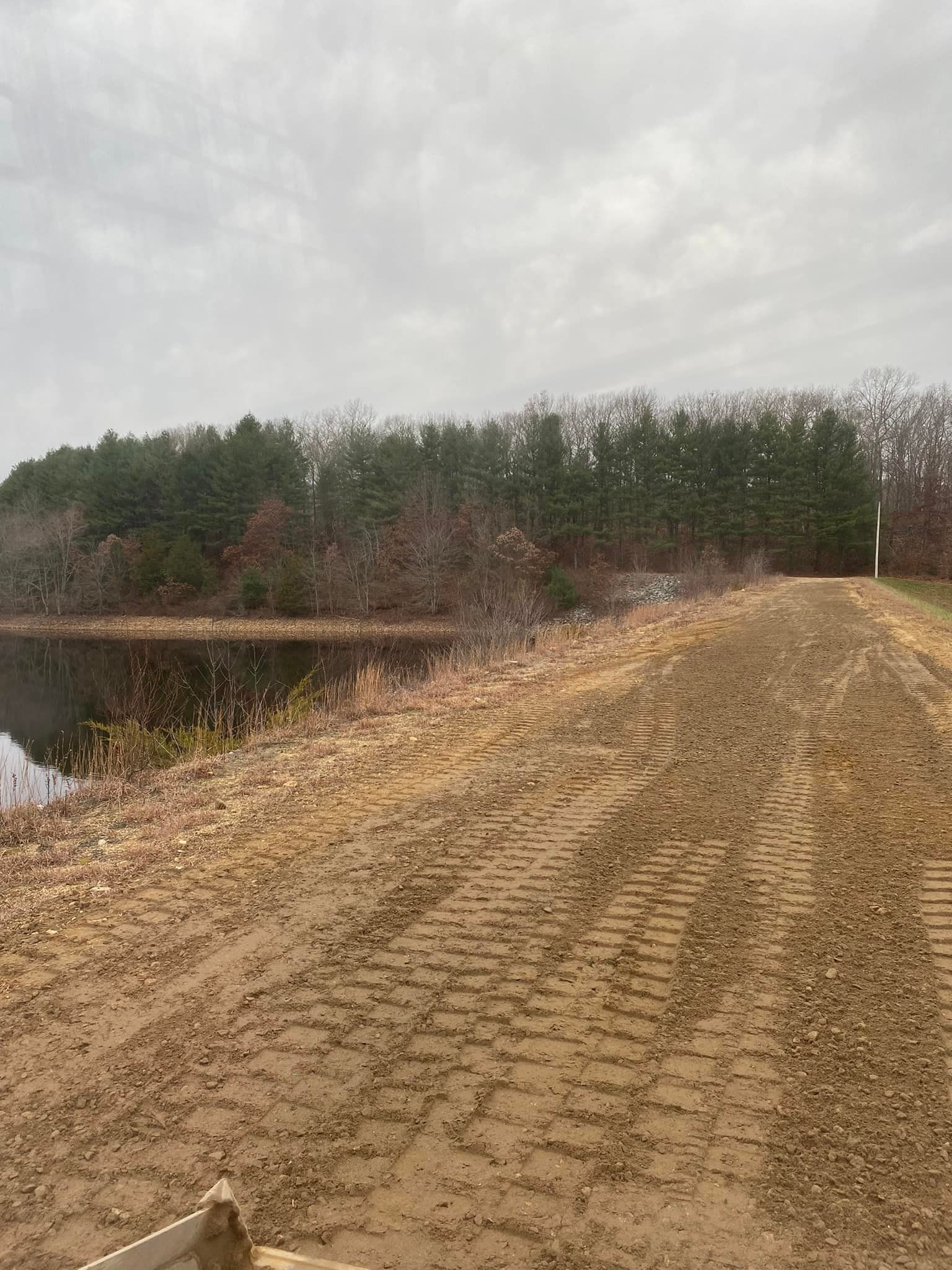 Dirt path alongside a body of water, leading toward a treeline under a cloudy sky.