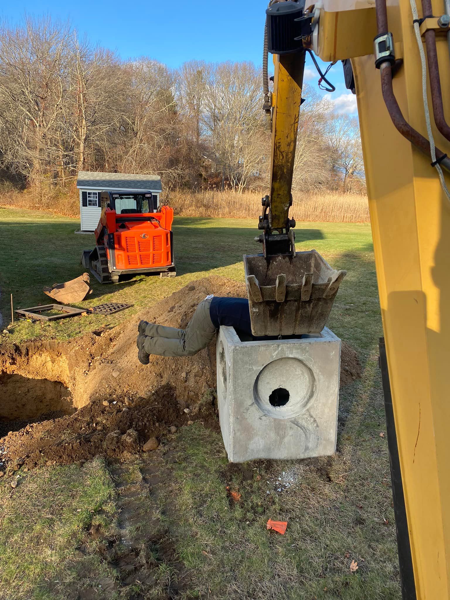 Person leaning into concrete block, held by excavator. Construction site with orange skid steer in background.