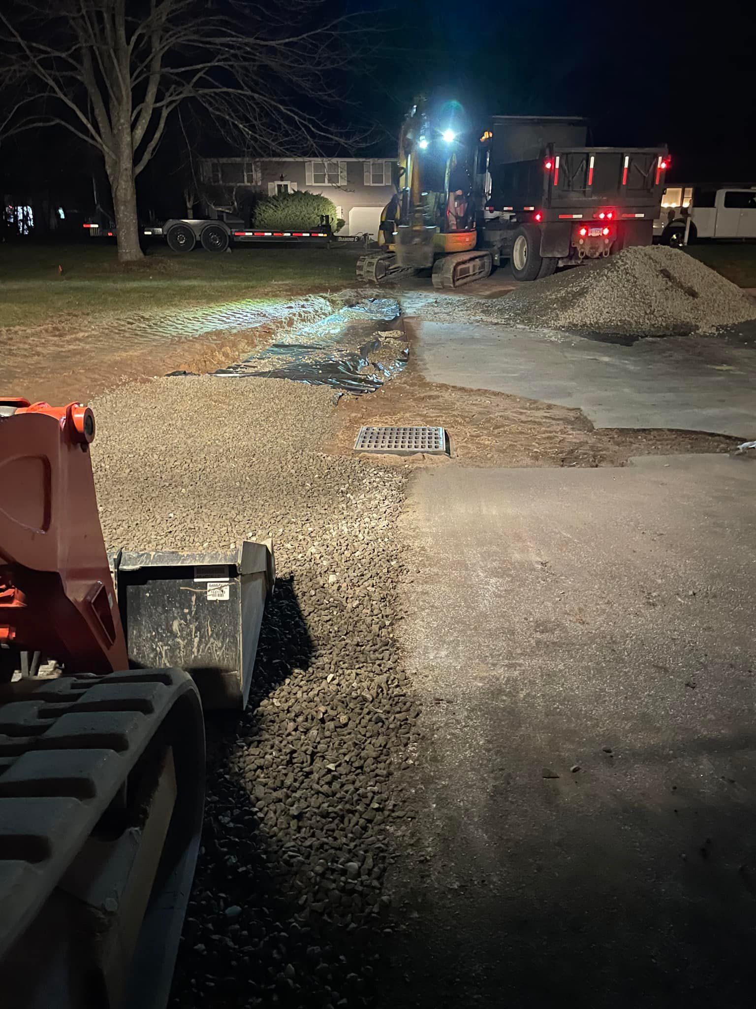 Construction site at night. Excavator loads gravel into a dump truck, with a skid steer in the foreground.