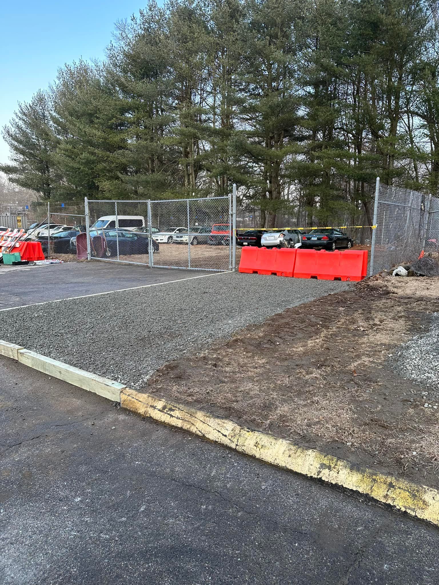 Gravel and dirt area next to a paved road, leading to a parking lot. Red barriers and a chain-link fence are present.
