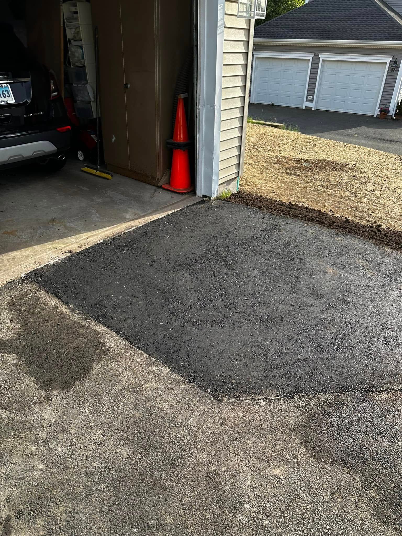Patch of fresh asphalt next to a garage, contrasting with gravel driveway. Orange cones visible.