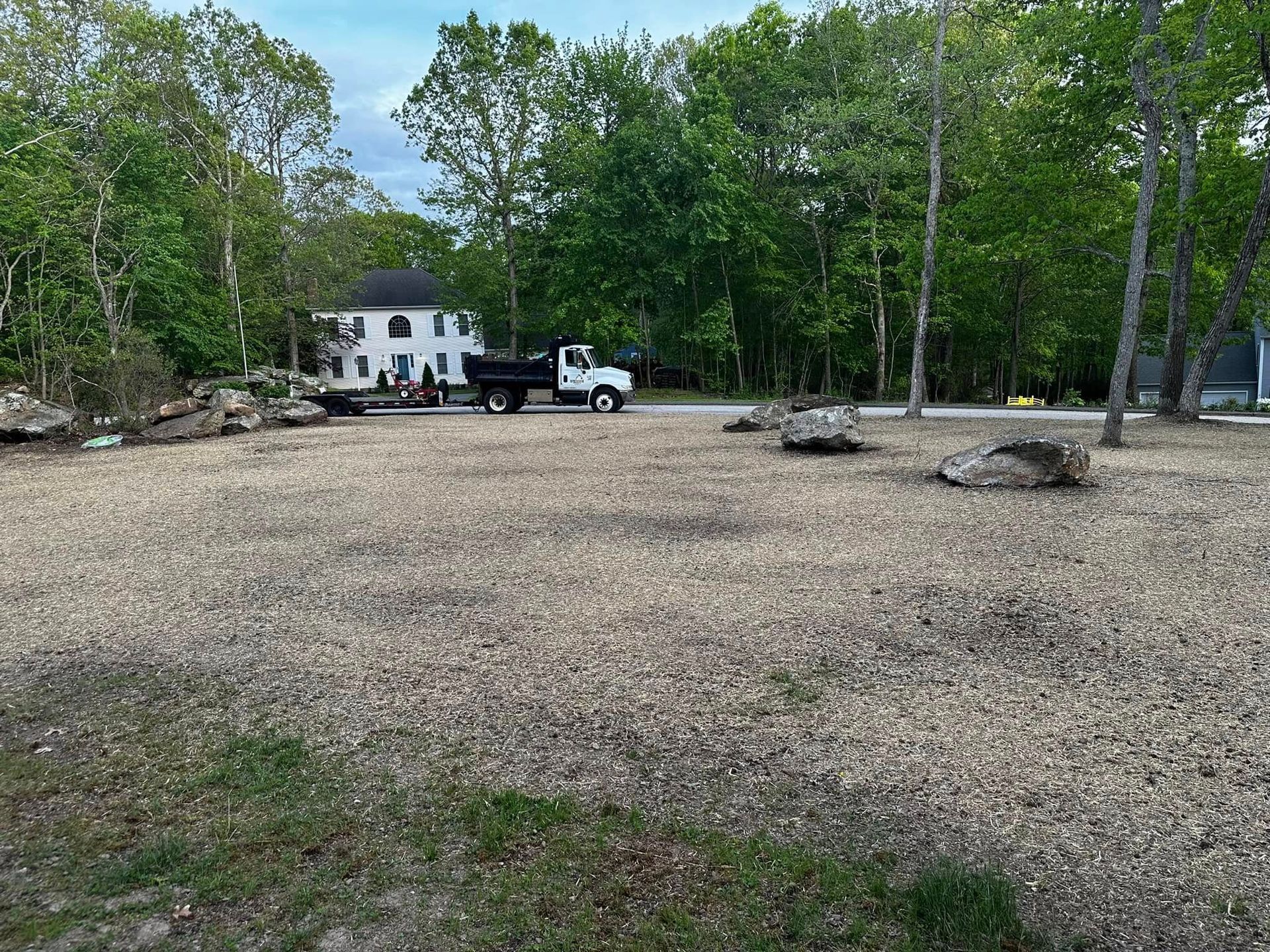Gravel lot with a white truck, house, and trees. Large rocks are scattered throughout the lot.