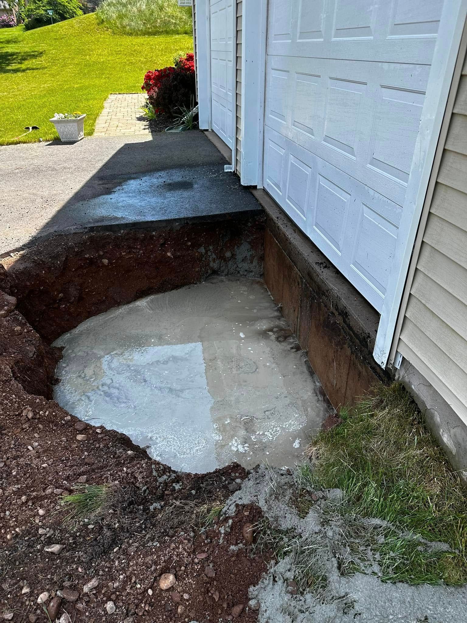 A hole filled with wet concrete beside a white garage. Dirt and grass surround it.