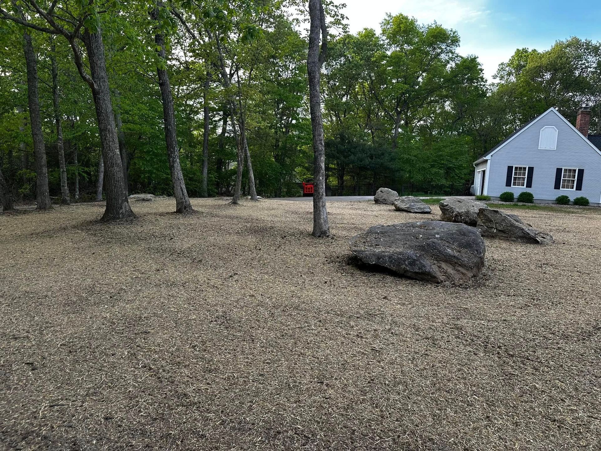 Gravel yard with large rocks, several trees, and a small, striped-sided building with a peaked roof.