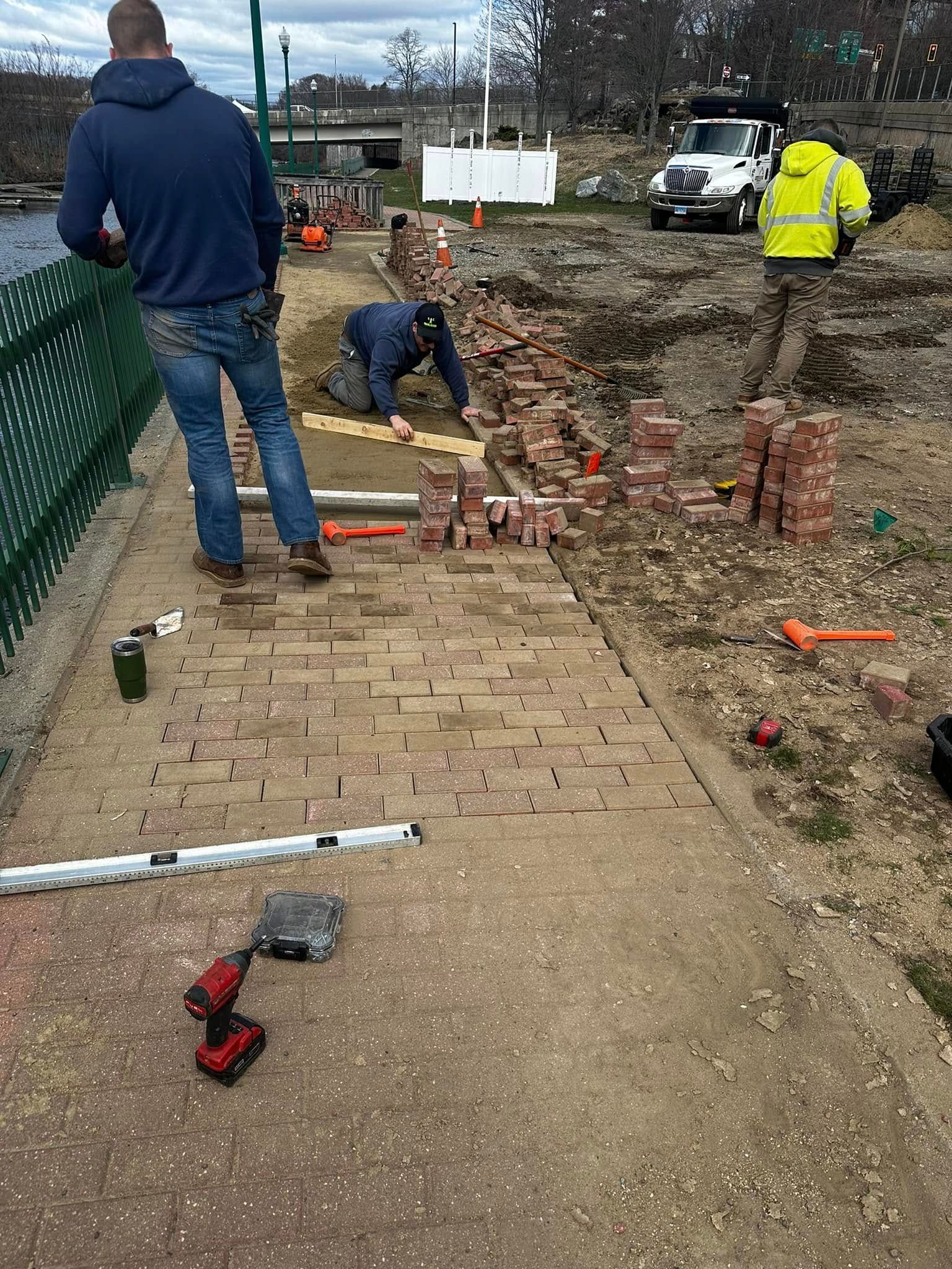 Construction workers laying bricks on a pathway by a waterway.