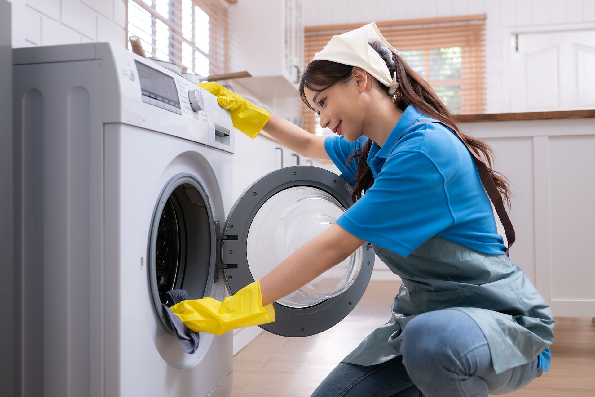 Woman wearing gloves and apron loading laundry into a washing machine in a brightly lit kitchen.