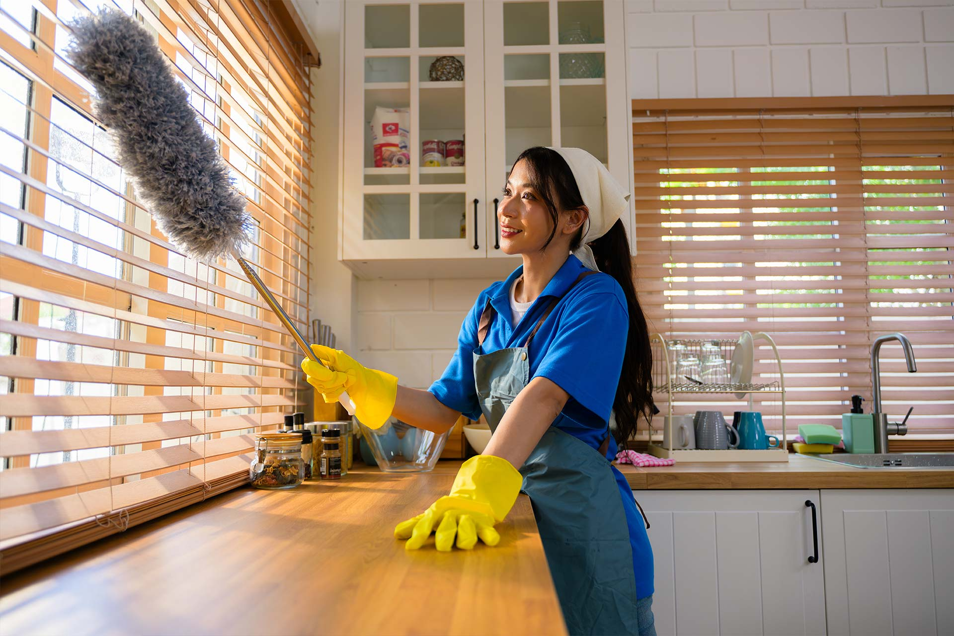 Woman dusting blinds in a kitchen, wearing yellow gloves and a blue uniform, sunlight shining.