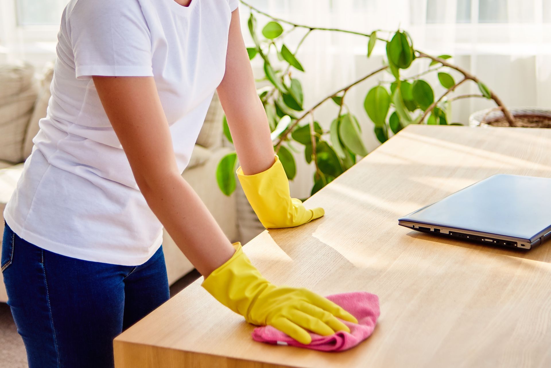 Woman in yellow gloves cleaning a wooden table with a pink cloth; laptop sits on the table.
