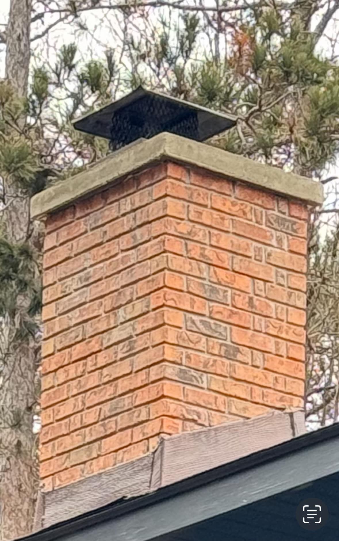 A brick chimney is sitting on top of a roof next to a tree.