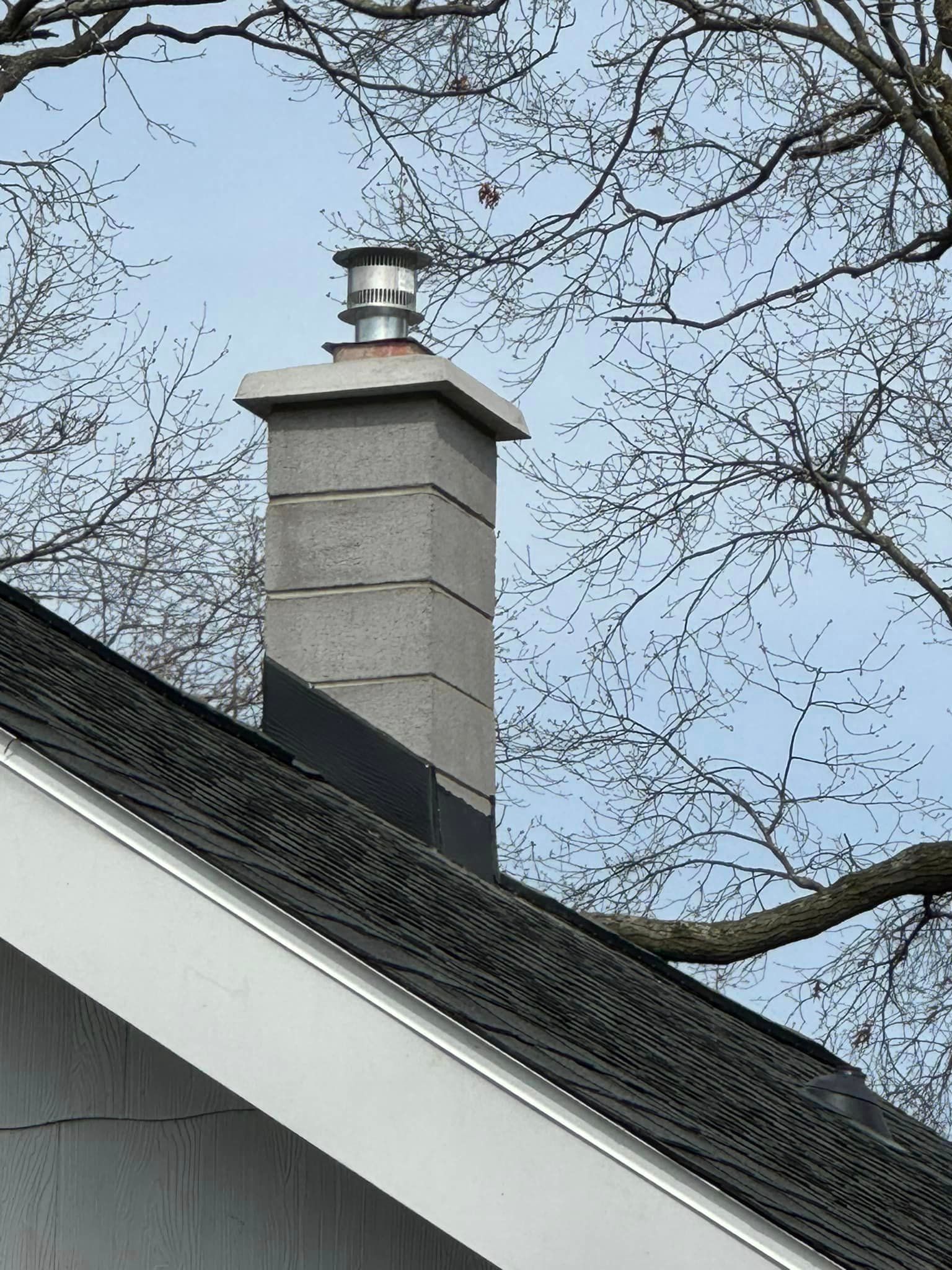 Chimney made of gray blocks on a dark shingled roof, topped with a metal cap, branches in the background.