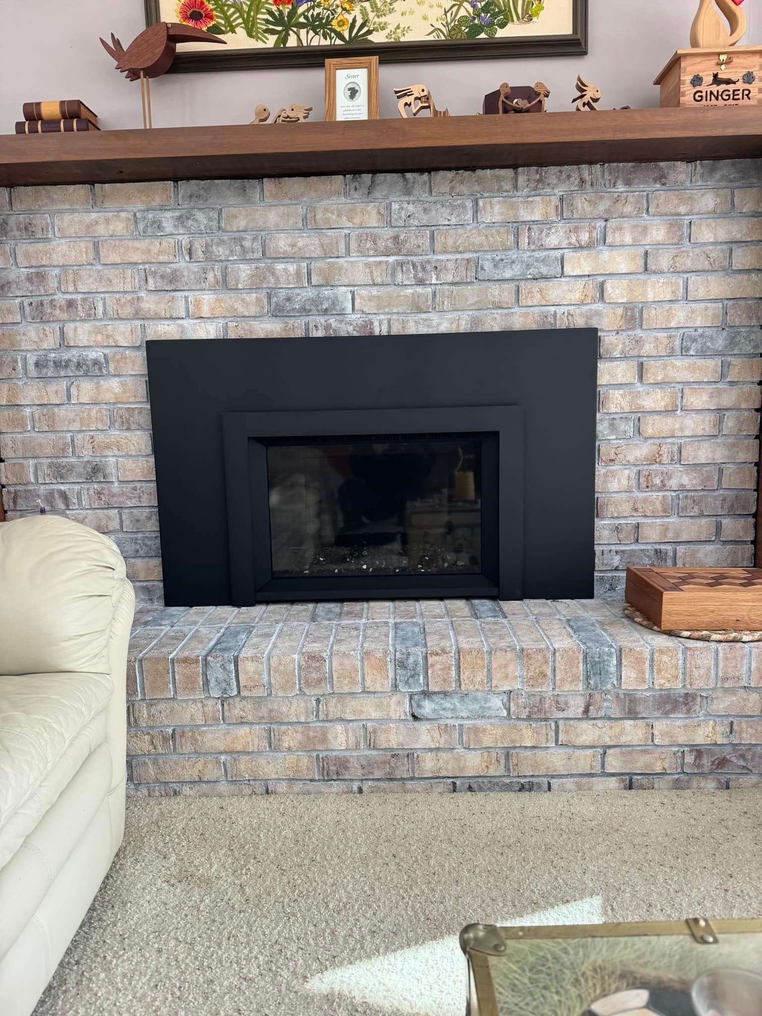 Brick fireplace with black surround; beige couch on left; wooden mantel with decor.