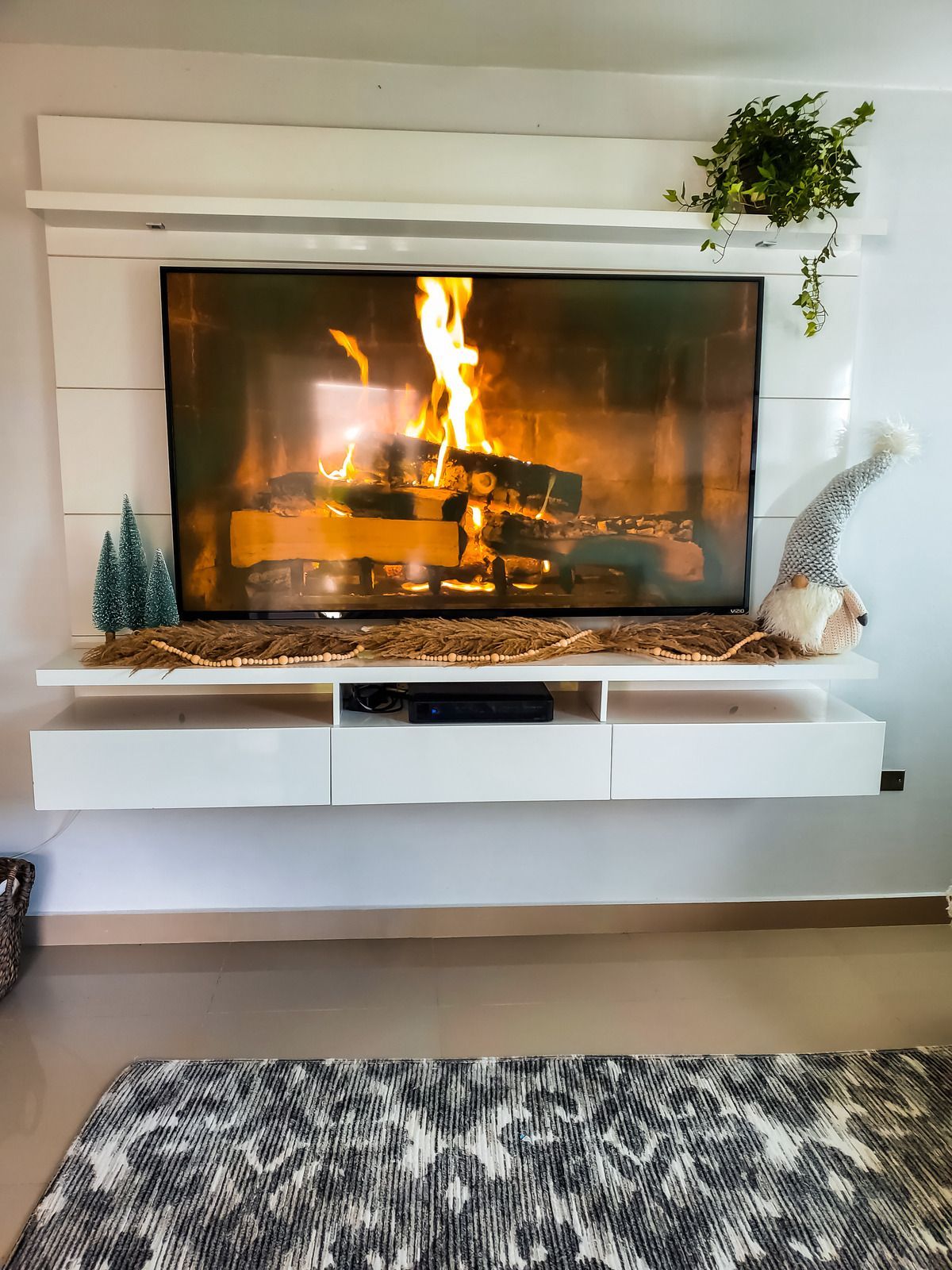 A TV displaying a fireplace scene, mounted on a white wall unit with decor, above a patterned rug.