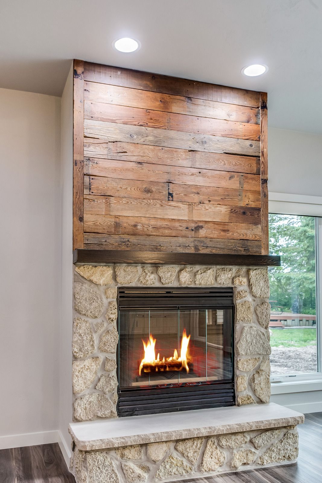 Fireplace with stone facade, wood mantel, and rustic wood paneling. Flames visible, set in a bright room.