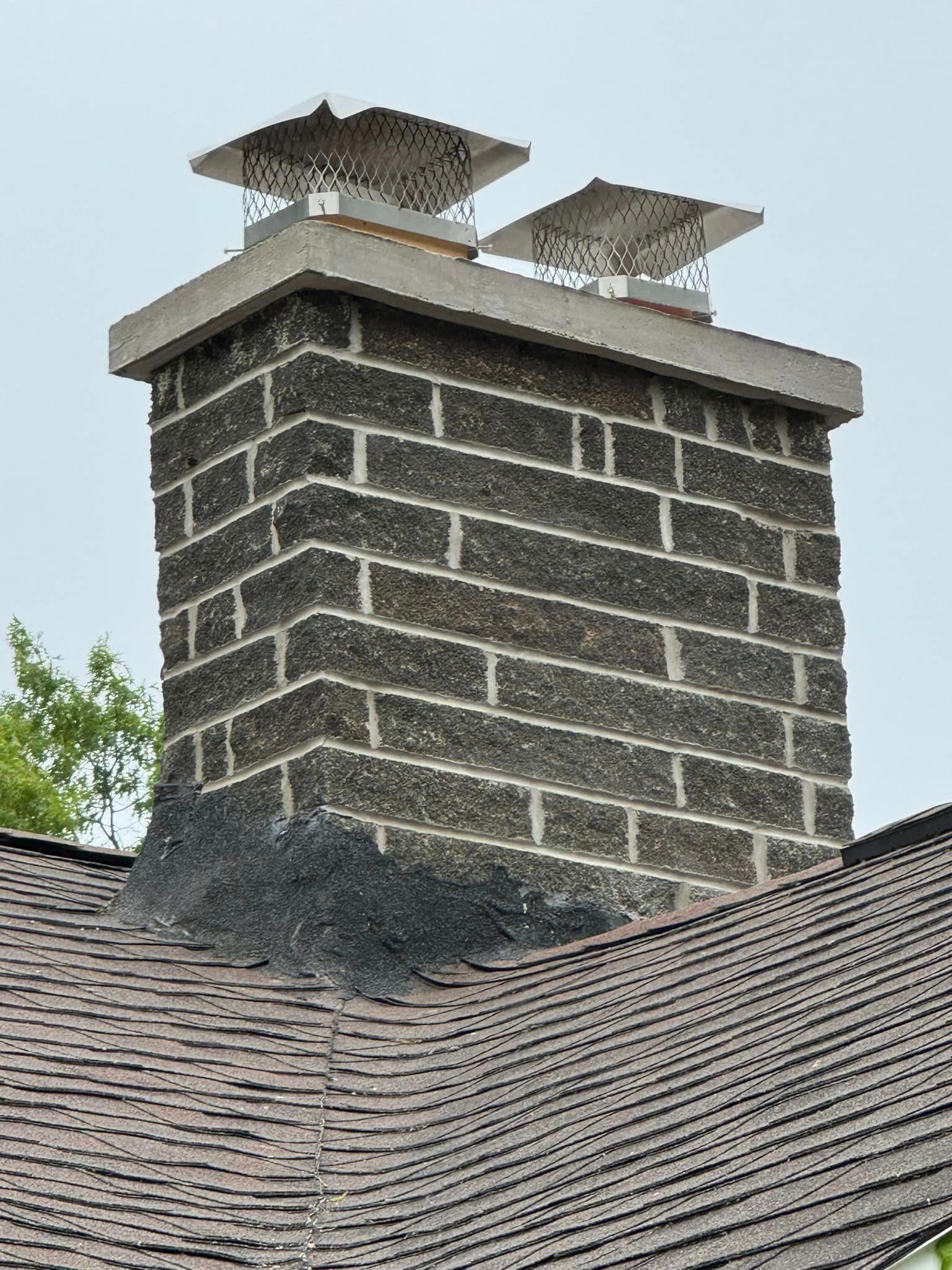 Brick chimney with concrete cap and metal chimney caps on a brown shingled roof.
