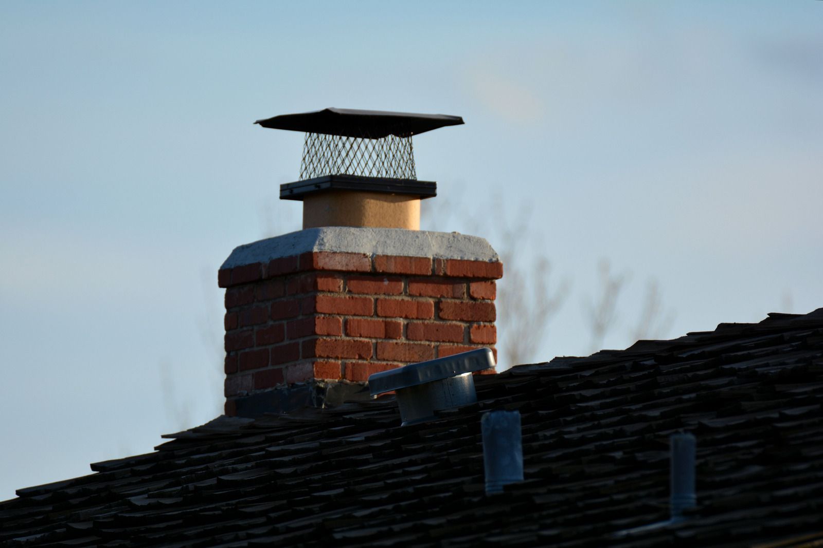 Brick chimney with a metal cap on a dark tiled roof against a blue sky.