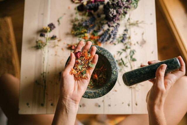 Hands grinding colorful dried herbs in a dark mortar and pestle on a wooden table.