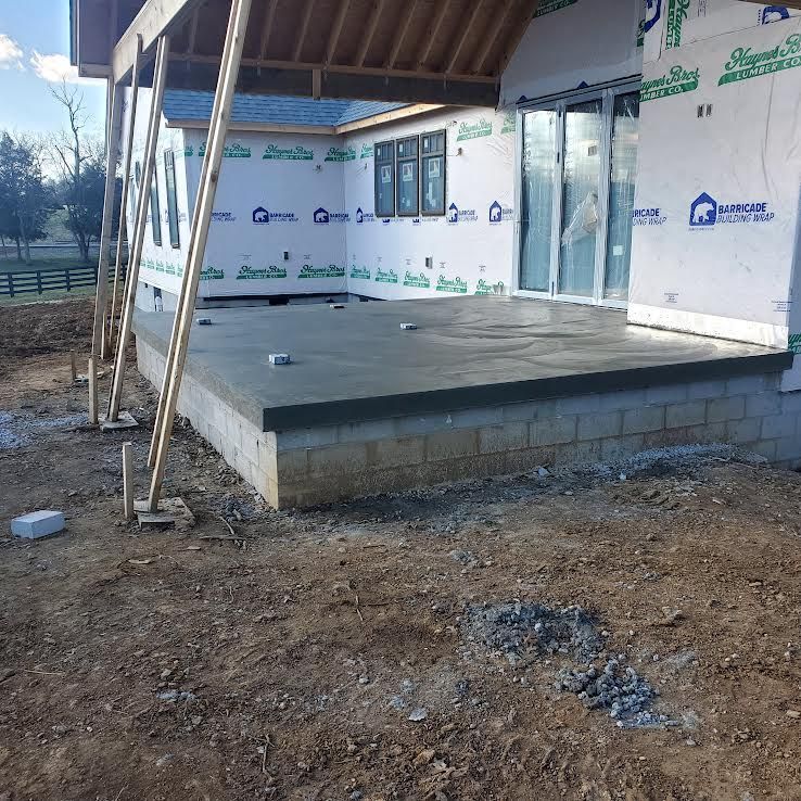 Concrete patio under construction with wooden supports, next to a house wrapped in blue barrier.