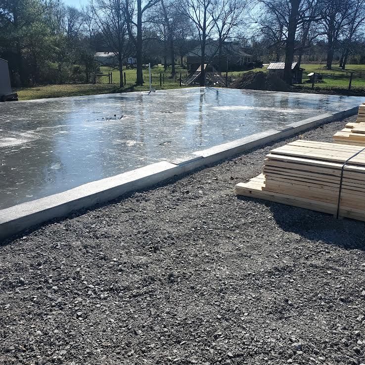 Concrete foundation with a gravel base, lumber stacked nearby, outdoors in a sunny setting.