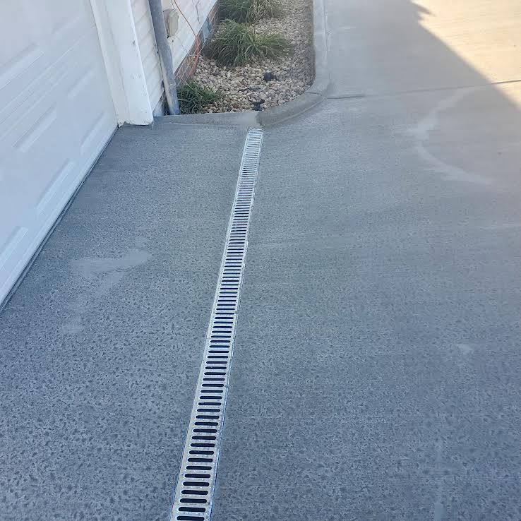 Concrete driveway with a linear drain, next to a garage door and landscaping.
