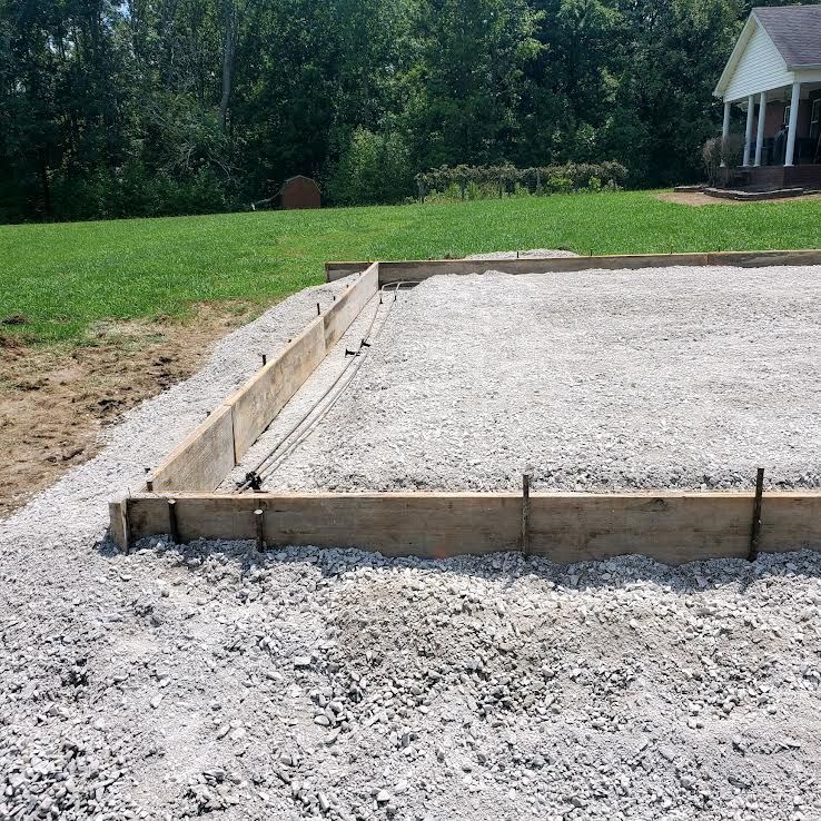 Prepared concrete foundation with wooden frame, set on gravel. Backyard setting, sunlight.