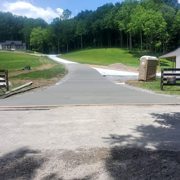 A new concrete driveway leads toward a house, with a porta-potty and a grassy hill in the background.