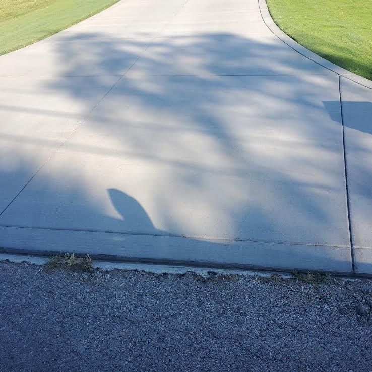 Concrete driveway curves to the right, with shadow from tree overhead. Bordered by grass and gravel.