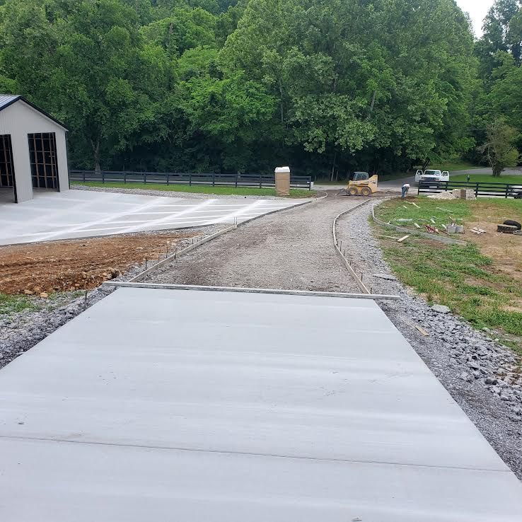 Construction site with a concrete driveway leading to a building and gravel path. Trees in the background.